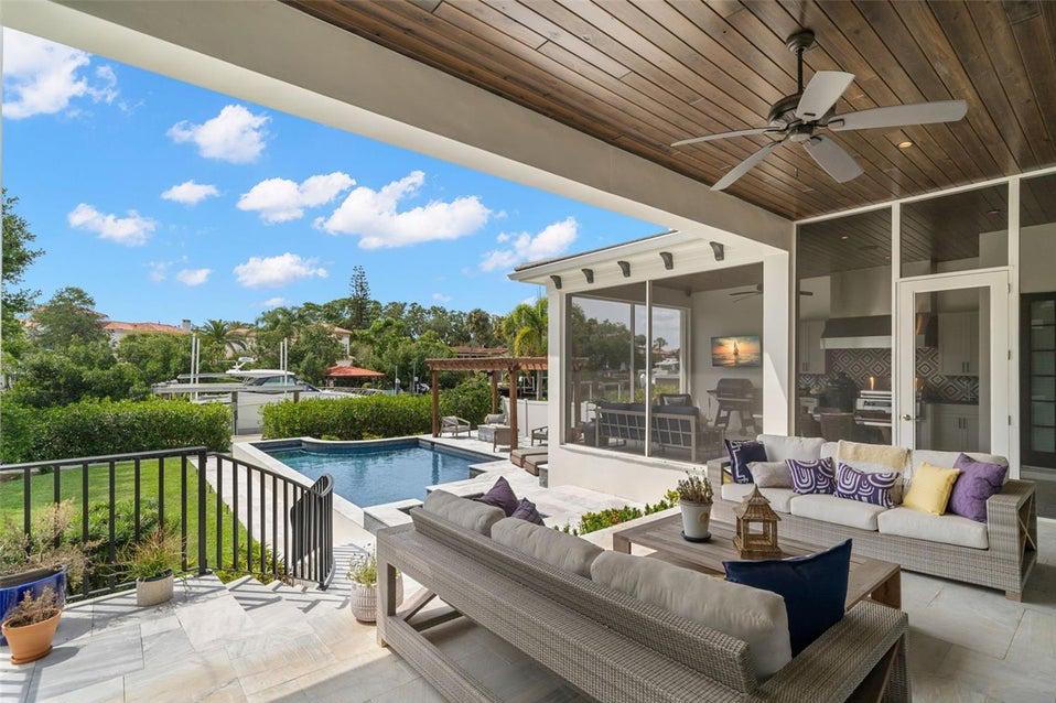 Outdoor patio with beige wicker furniture, a pool, and a covered deck connected to a glass-walled home interior on a sunny day.