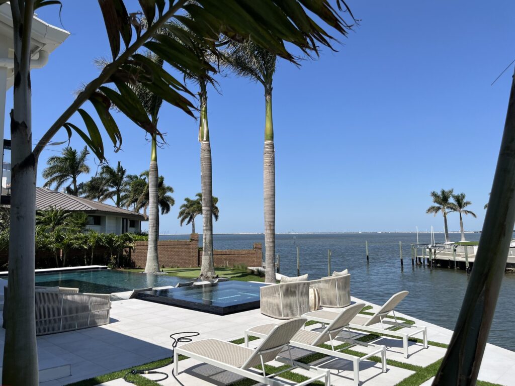 Poolside patio with white lounge chairs, palm trees, and a blue water canal under a clear sky.