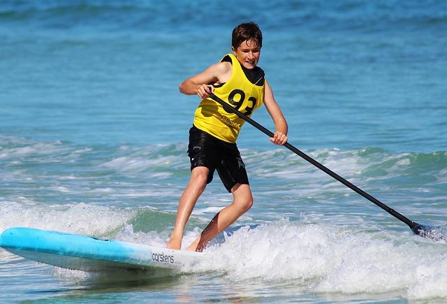 Boy wearing a yellow race vest paddling a blue stand-up paddleboard on the ocean waves near shore.