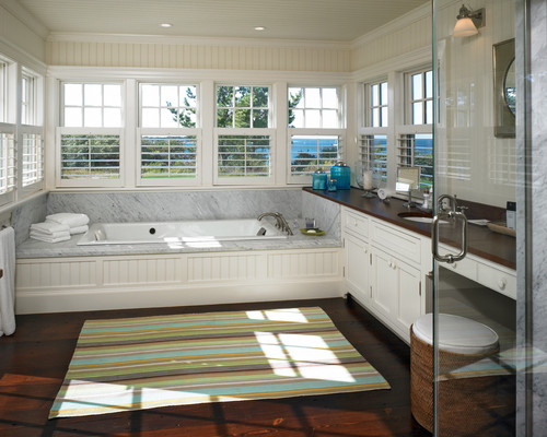 Bright sunlit bathroom with a white marble tub by large windows and white cabinetry, striped rug on dark wood floor.