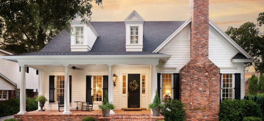 Front view of a white two-story house with a covered porch, rocking chairs, and a tall brick chimney at dusk.